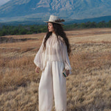 A woman with long dark hair, wearing a white wide-brimmed hat and AYA Sacred Wear’s Apsara Pants in cream—pure silk flowy pants—stands in a dry grassy field before a mountain, gazing to the side and holding a small book under a partly cloudy sky.