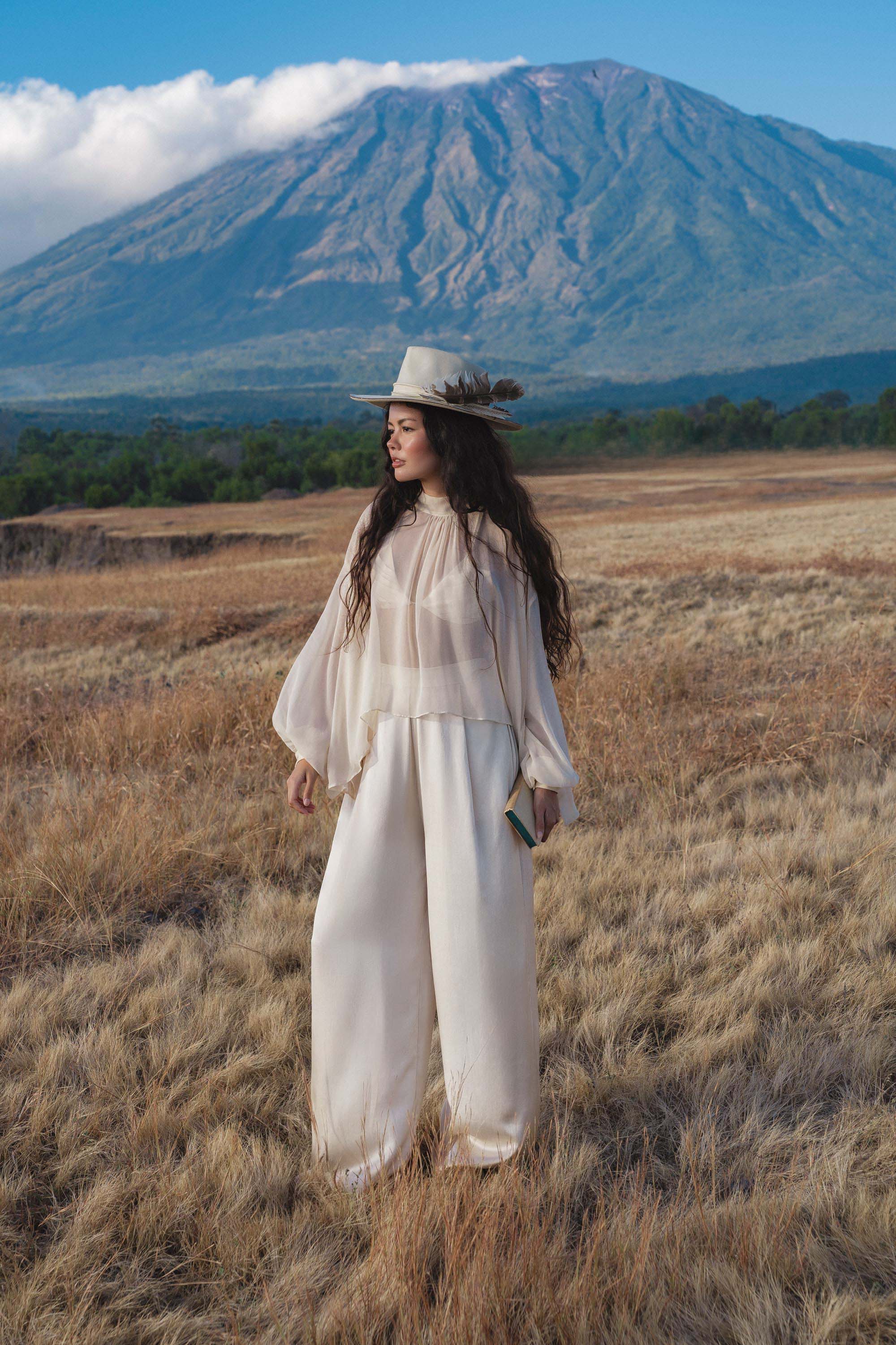 A woman with long dark hair, wearing a white wide-brimmed hat and AYA Sacred Wear’s Apsara Pants in cream—pure silk flowy pants—stands in a dry grassy field before a mountain, gazing to the side and holding a small book under a partly cloudy sky.