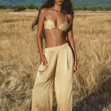 A woman with long dark hair stands in a dry grassy field, wearing a beige bralette and AYA Sacred Wear’s Apsara Pants Inka Gold – Pure Silk Flowy Pants. Mountains and clouds form the backdrop as she gazes confidently at the camera, one hand in her pocket.