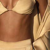 A person wears AYA Sacred Wear's Apsara Pants Inka Gold—pure silk, flowy pants—in light gold, paired with a silky bralette and draped fabric on the arm. The softly blurred background creates an elegant outdoor vibe.