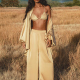 A woman with long wavy hair stands in a dry grass field wearing AYA Sacred Wear's Apsara Pants Inka Gold—pure silk flowy pants inspired by handcrafted Bali pants. Mountains and greenery appear in the distance under a clear sky.