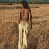 A woman with long dark hair, wearing a beige backless strappy top and AYA Sacred Wear’s Apsara Pants Inka Gold – Pure Silk Flowy Pants, walks through tall golden grass in a field with mountains and trees in the background, facing away from the camera.