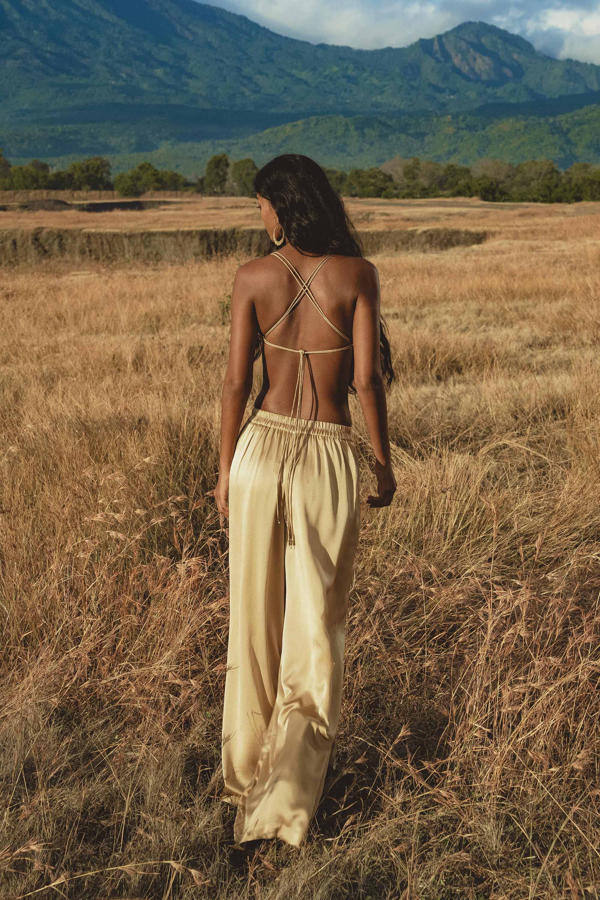 A woman with long dark hair, wearing a beige backless strappy top and AYA Sacred Wear’s Apsara Pants Inka Gold – Pure Silk Flowy Pants, walks through tall golden grass in a field with mountains and trees in the background, facing away from the camera.