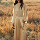 A woman with long wavy hair stands in a sunlit, grassy field wearing AYA Sacred Wear’s Apsara Pants Inka Gold—pure silk flowy pants—and a sheer gold blouse, holding a camera as scattered trees bask in the soft light.