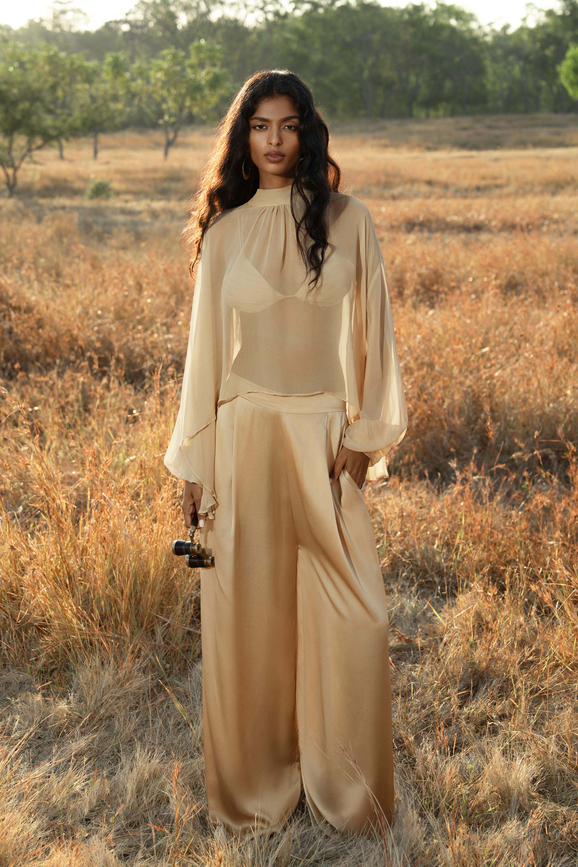 A woman with long wavy hair stands in a sunlit, grassy field wearing AYA Sacred Wear’s Apsara Pants Inka Gold—pure silk flowy pants—and a sheer gold blouse, holding a camera as scattered trees bask in the soft light.