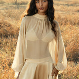 A woman with long wavy hair stands outdoors in a sunlit field, wearing AYA Sacred Wear's Apsara Pants Inka Gold—pure silk flowy pants—with a light beige blouse and matching top, calm as she holds a small object amid trees and dry grass.