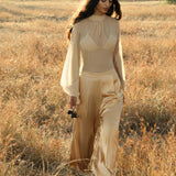 A woman with long, wavy hair strolls through a sunlit field in a sheer beige top and AYA Sacred Wear’s Apsara Pants Inka Gold – pure silk flowy pants, looking down at her camera amid the serene natural beauty.