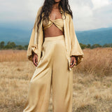 A woman with long, wavy dark hair stands in a dry grassy field wearing AYA Sacred Wear's Apsara Pants Inka Gold—pure silk flowy pants—paired with a gold satin knotted crop top and oversized shirt, mountains and clouds behind her as she gazes confidently at the camera.