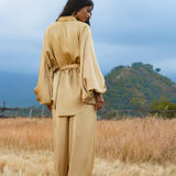 A woman stands in a golden field, wearing AYA Sacred Wear’s Apsara Pants Inka Gold—pure silk flowy pants. Her hair is down as she faces away, with tree-covered hills and a cloudy sky in the background.