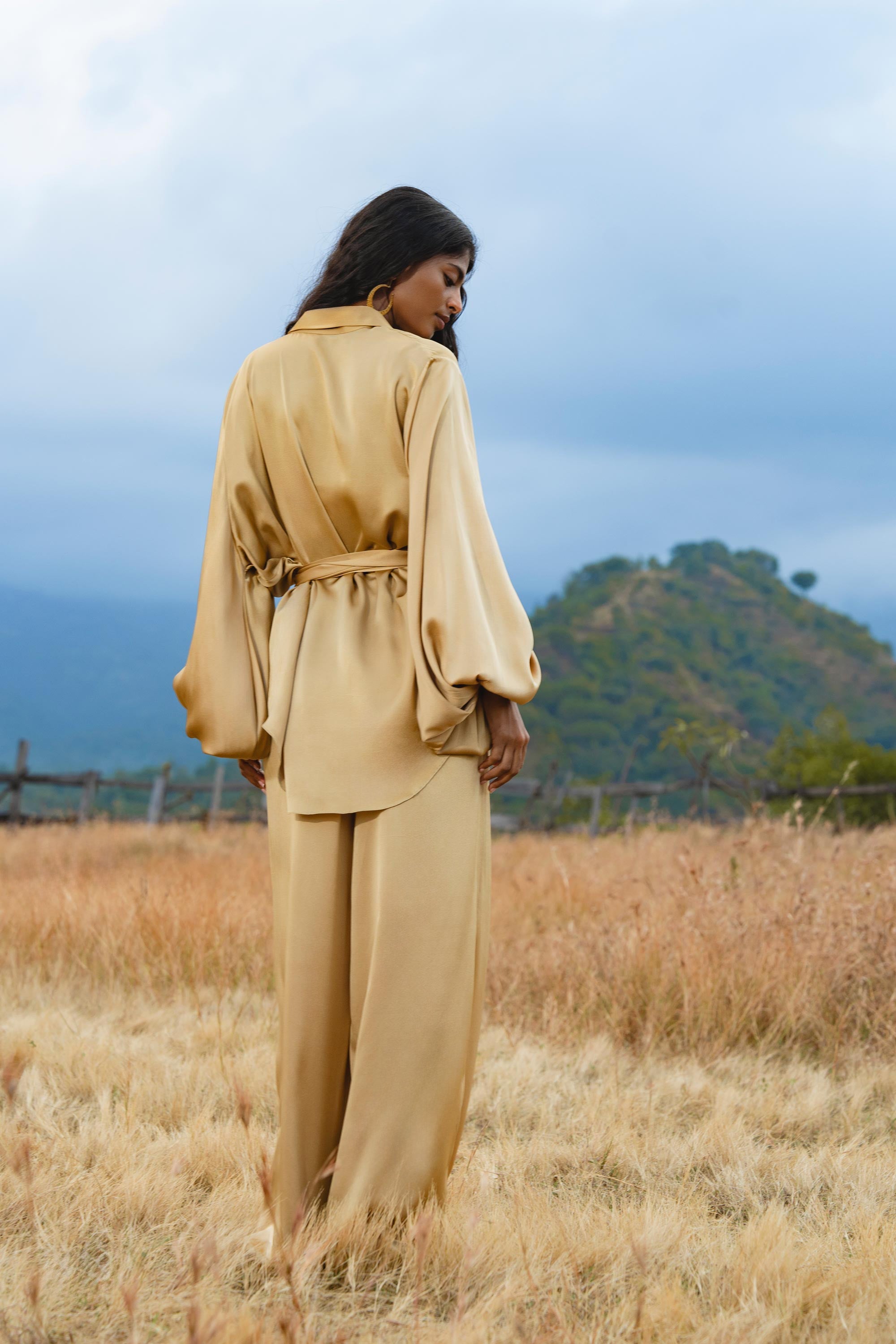 A woman stands in a golden field, wearing AYA Sacred Wear’s Apsara Pants Inka Gold—pure silk flowy pants. Her hair is down as she faces away, with tree-covered hills and a cloudy sky in the background.