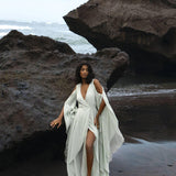 On a rocky beach, a woman in an AYA Sacred Wear Aquamarine Sundari Goddess Silk Gown, featuring ahimsa silk, a deep V-neck, and slit stands barefoot against dark rocks with waves in the background under an overcast sky.