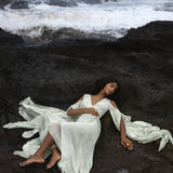 A woman in a Sky Blue Makosh Goddess Dress by AYA Sacred Wear lies gracefully on dark, rocky terrain near an ocean with waves crashing. The serene and dramatic scene highlights the contrast between the soft, light cotton wrap dress and the rugged landscape.