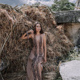 A woman wearing a Beige Organic Cotton Bohemian Tribal Maxi Dress with Hand Loomed Tassels from AYA Sacred Wear sits barefoot on stone steps in front of a large stack of hay. The sky above is partly cloudy, and trees can be seen in the background.