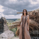 A woman stands barefoot in front of a rustic landscape, showcasing the Beige Organic Cotton Bohemian Tribal Maxi Dress with Hand Loomed Tassels by AYA Sacred Wear. The background features dramatic clouds and piles of dried grass.