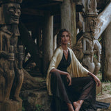 A woman wearing a Black Boho Jumpsuit by AYA Sacred Wear, featuring handmade cold shoulder details, sits barefoot on a stone step near intricately carved wooden statues under a rustic wooden structure. The setting is enveloped in an ancient, tribal vibe highlighted by natural earth tones and textures.