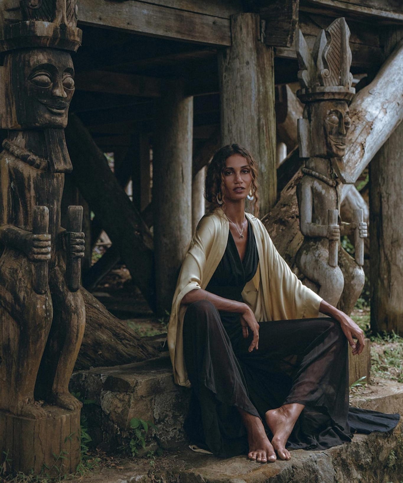 A woman wearing a Black Boho Jumpsuit by AYA Sacred Wear, featuring handmade cold shoulder details, sits barefoot on a stone step near intricately carved wooden statues under a rustic wooden structure. The setting is enveloped in an ancient, tribal vibe highlighted by natural earth tones and textures.
