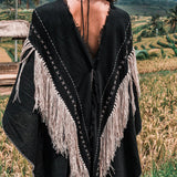A woman with long dark hair stands in a field, enveloped in the AYA Sacred Wear Black Boho Poncho, adorned with hand stitching and hand loomed tassels. The background features terraced greenery and palm trees beneath a cloudy sky.