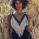 A person with curly hair stands outdoors amidst tall grasses, wearing the Black Boho Tunic Top by AYA Sacred Wear, featuring tassels and hand embroidery. The sunlight highlights the natural scenery while beautifully complementing the hand-loomed cotton fabric's texture.
