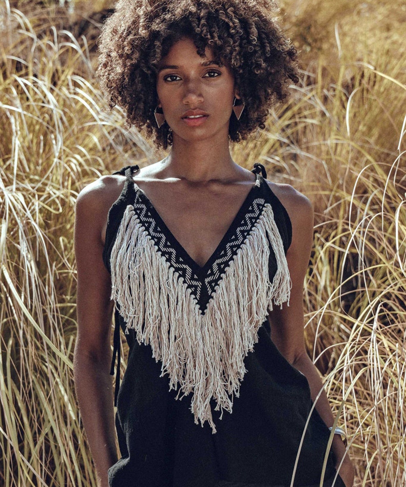 A person with curly hair stands outdoors amidst tall grasses, wearing the Black Boho Tunic Top by AYA Sacred Wear, featuring tassels and hand embroidery. The sunlight highlights the natural scenery while beautifully complementing the hand-loomed cotton fabric's texture.