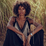 Amidst the tall grass, a person with curly hair sits serenely, dressed in a black outfit featuring intricate front detailing and a silver bracelet. The sunlight highlights their calm expression, while the Black Boho Tunic Top with Tassels and Hand Embroidery by AYA Sacred Wear, made from hand-loomed cotton, adds an earthy touch to the scene.