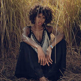 A person with curly hair sits among tall, dry grass, sporting a sleeveless Black Boho Tunic Top with Tassels and Hand Embroidery from AYA Sacred Wear and black pants. They have a thoughtful expression and a silver bracelet on their right wrist, surrounded by a natural and warm setting.