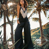 A woman stands confidently among palm trees, showcasing the AYA Sacred Wear's Black Boho Tunic Top with Tassels and Hand Embroidery paired with wide-legged pants. The sun streams through the leaves, crafting a tropical backdrop with hills in the distance, perfectly embodying a vibe of sustainable fashion.