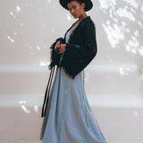 A person poses outdoors in a stylish outfit, featuring a wide-brimmed black hat, the Black Boho Wrap Top from AYA Sacred Wear with its durable fringe details and wide sleeves, and a long light blue dress. The background is softly lit with dappled shadows from nearby foliage.