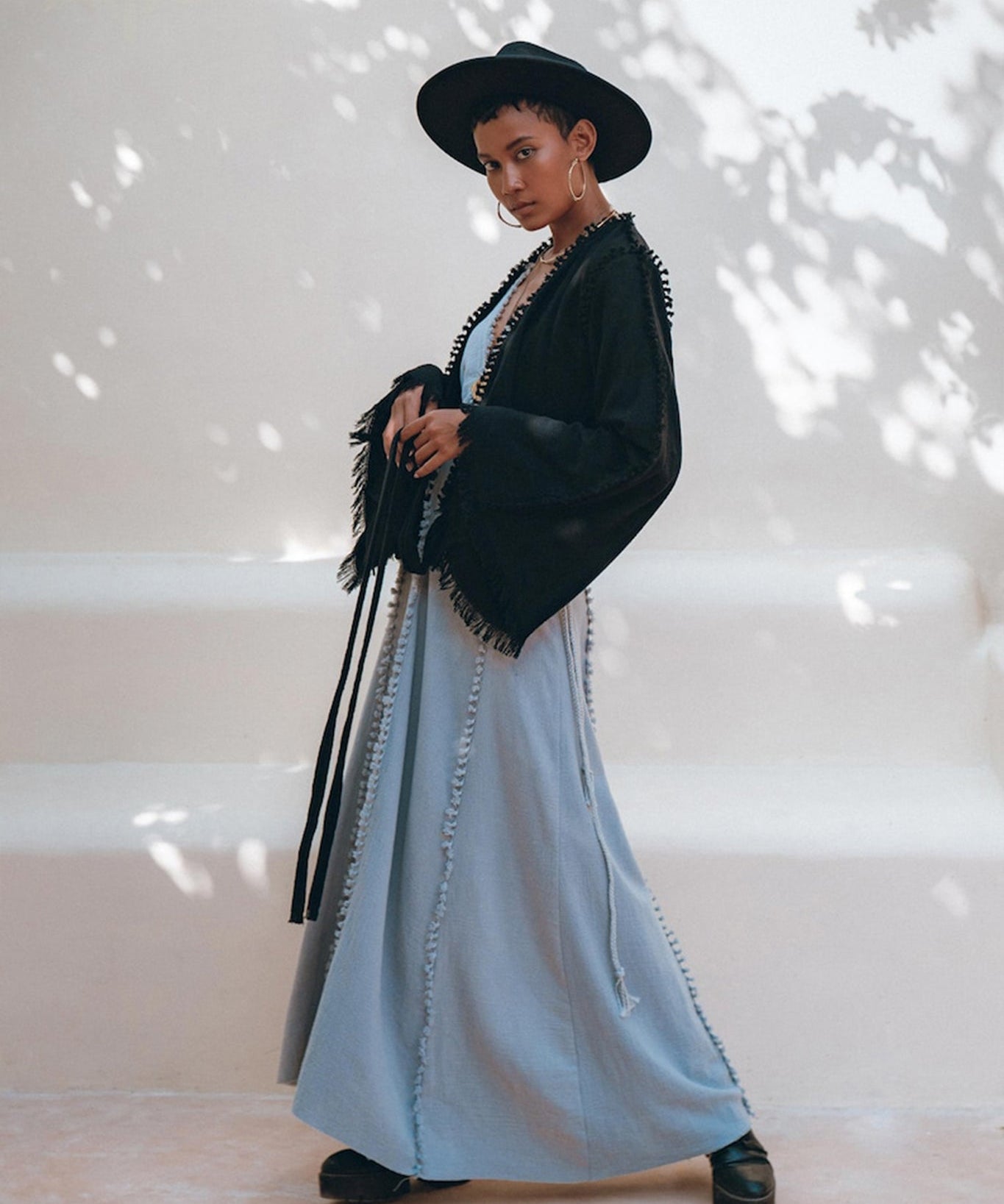 A person poses outdoors in a stylish outfit, featuring a wide-brimmed black hat, the Black Boho Wrap Top from AYA Sacred Wear with its durable fringe details and wide sleeves, and a long light blue dress. The background is softly lit with dappled shadows from nearby foliage.