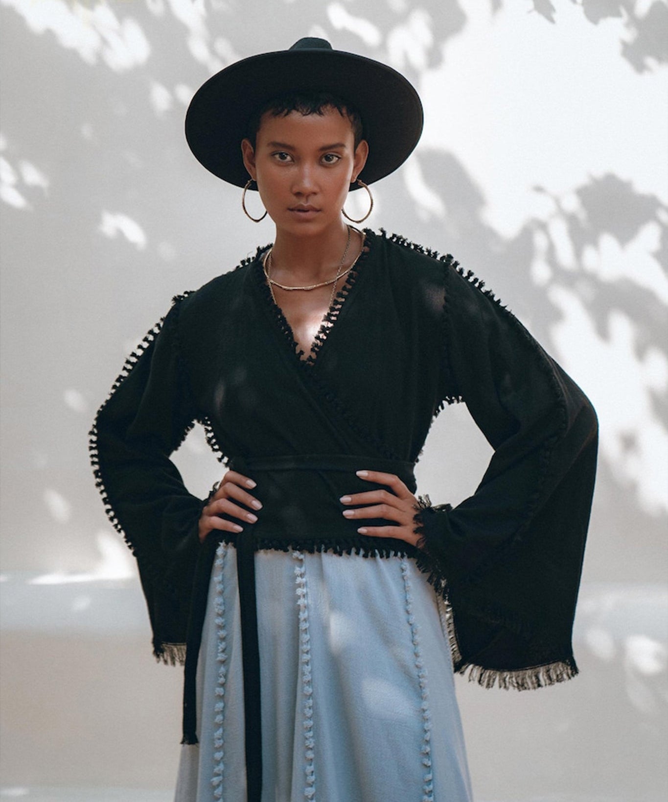 A fashionista poses confidently outdoors, wearing a black wide-brimmed hat and the Black Boho Wrap Top with wide sleeves by AYA Sacred Wear. Paired with a gray skirt featuring decorative details, her ensemble shines in the dappled sunlight, while tree shadow patterns on the wall behind enhance the scene.