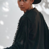 A person wearing a Black Boho Wrap Top with wide sleeves from AYA Sacred Wear and a black textured jacket, evoking the essence of a durable garment, looks back over their shoulder. The background is softly blurred with the gentle outline of tree shadows.