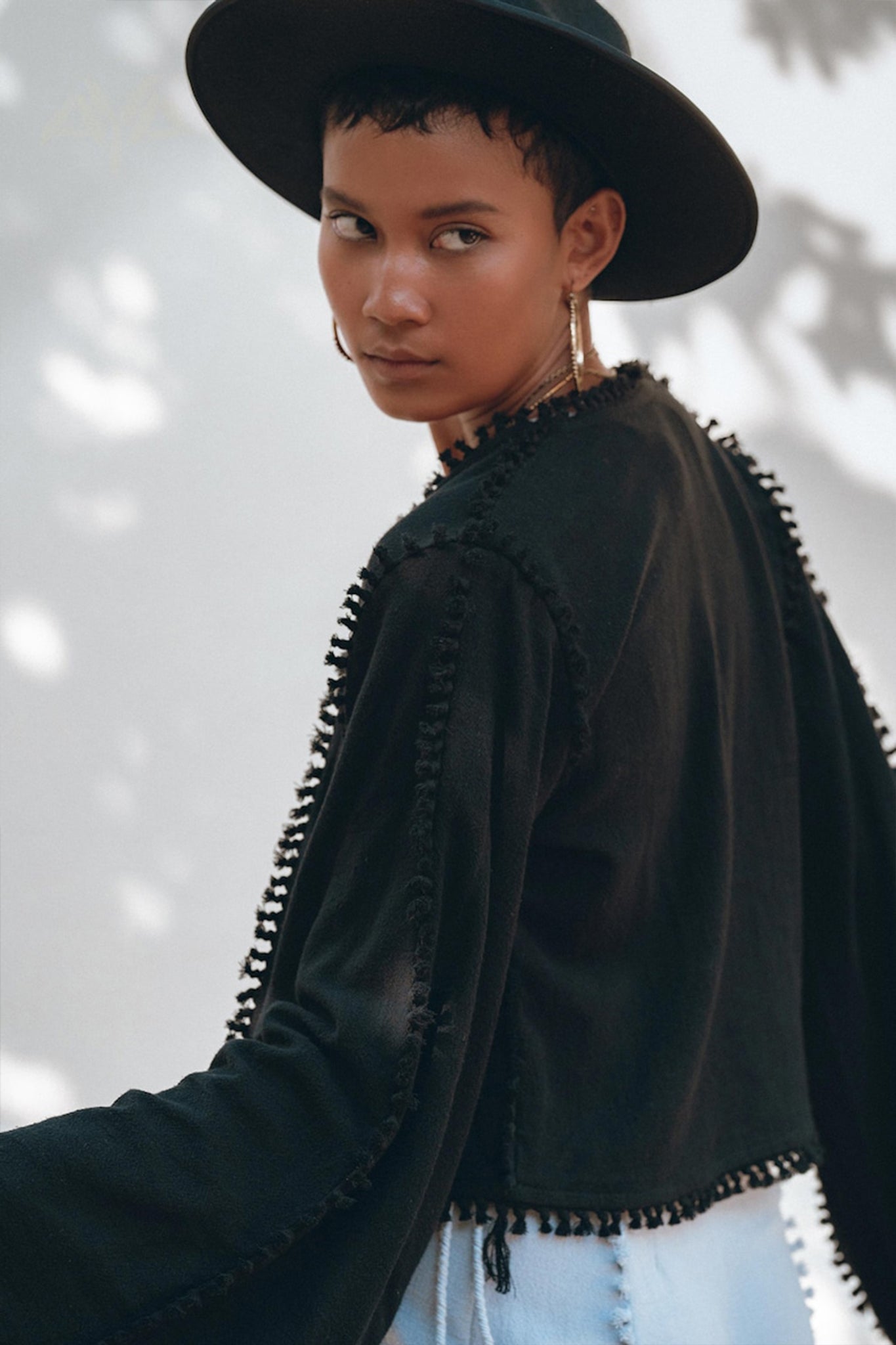 A person wearing a Black Boho Wrap Top with wide sleeves from AYA Sacred Wear and a black textured jacket, evoking the essence of a durable garment, looks back over their shoulder. The background is softly blurred with the gentle outline of tree shadows.