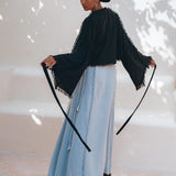An individual wearing the AYA Sacred Wear's Black Boho Wrap Top, featuring wide sleeves and fringe details, paired with a light blue organic cotton skirt, stands facing backward while tying a knot. The softly lit background creates a captivating shadow effect, allowing their handmade design to shine through the ensemble.