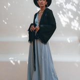 A person stands confidently wearing a black wide-brimmed hat, large hoop earrings, and a black fringed jacket. They pair this ensemble with the Black Boho Wrap Top from AYA Sacred Wear, crafted from organic cotton with decorative trim. The outfit is completed by a long light blue dress that adds an ethereal touch to the look. They pose against a neutral backdrop with soft shadows for an effortlessly chic appearance.