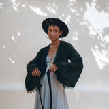 A woman wearing the Black Boho Wrap Top from AYA Sacred Wear, complemented by a wide-brimmed hat and a long light dress, stands against a sunlit wall with dappled shadows. The ensemble features a handcrafted design, with white flowers hanging above to create an artful scene.