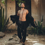 A bearded man stands outdoors in front of a decorative stone wall, wearing AYA Sacred Wear's Black Colour Handwoven Cotton Pants for Men. He pairs the pants with sandals and an open black shirt that reveals his chest adorned with a beaded necklace, surrounded by lush greenery and rocky ground.