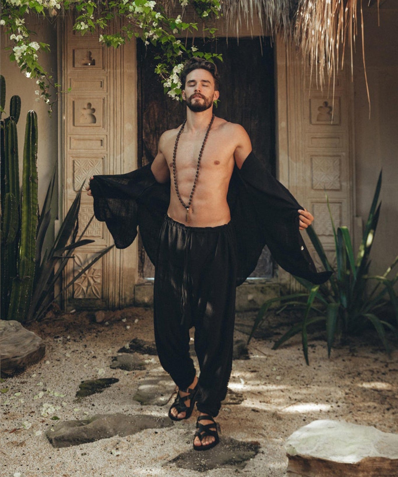 A bearded man stands outdoors in front of a decorative stone wall, wearing AYA Sacred Wear's Black Colour Handwoven Cotton Pants for Men. He pairs the pants with sandals and an open black shirt that reveals his chest adorned with a beaded necklace, surrounded by lush greenery and rocky ground.