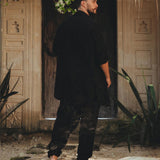 A person dressed in AYA Sacred Wear's Black Colour Handwoven Cotton Pants for Men stands on a sandy pathway before an ornate wooden door, enveloped by lush plants and greenery, enhancing the tranquil and natural ambiance of the scene.