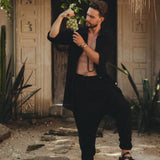 Outdoors in a tropical setting with lush greenery and an ornate, carved doorway in the background, a bearded man is holding a bunch of grapes. He is dressed in an open black shirt paired with Black Colour Handwoven Cotton Pants for Men by AYA Sacred Wear.