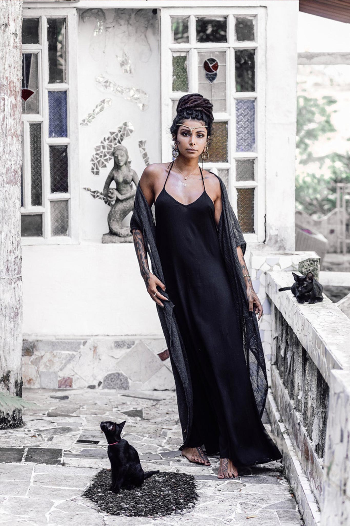 A woman in a Black Cotton Boho Wedding Slip Dress by AYA Sacred Wear stands on a stone terrace beside a black cat. Behind her is a wall adorned with stained glass windows and a statue. Her long dark hair is styled in an updo, she wears a necklace, and the cat sits at her feet.