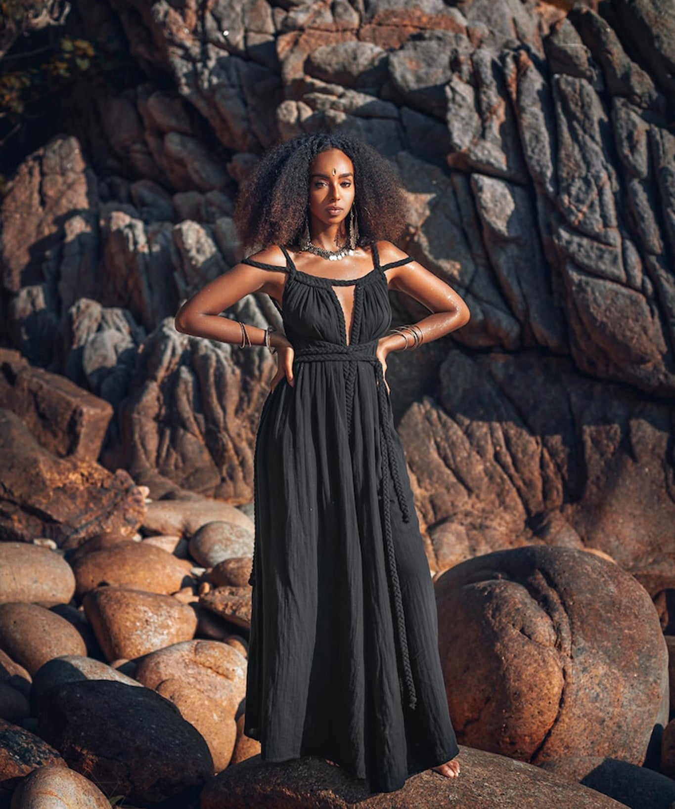 A woman stands confidently amidst rugged terrain, dressed in an elegant Black Greek Goddess Prom Boho Evening Dress from AYA Sacred Wear. Her hands rest on her hips, and her voluminous hair is beautifully highlighted by the warm, natural light against a backdrop of large stones and jagged rocks.