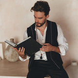 A man with dark hair and a beard is sitting on a ledge, engrossed in reading a book. He wears the Black Hand Embroidered Vest for Men from AYA Sacred Wear over his white shirt. Ceramic vases add elegance to the background against the textured beige wall.
