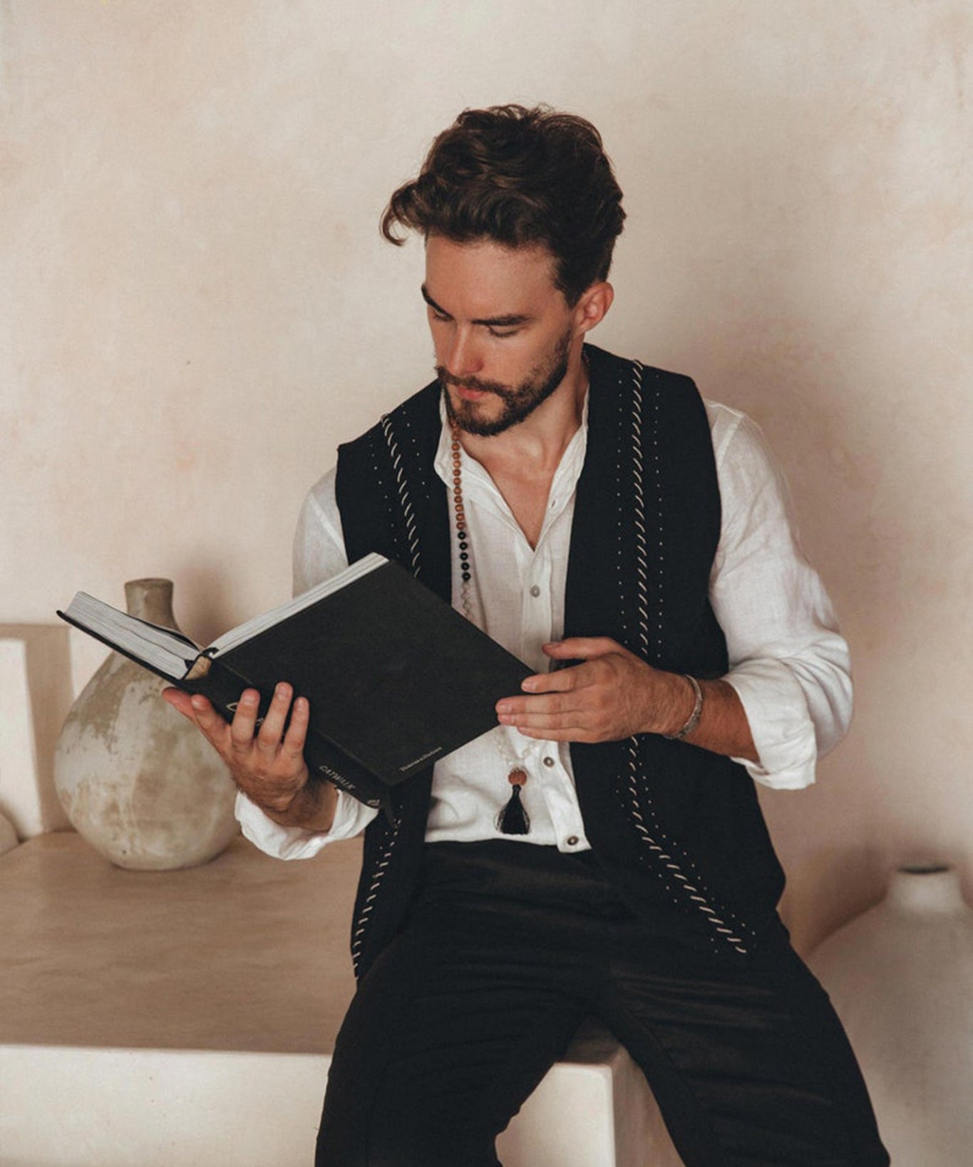 A man with dark hair and a beard is sitting on a ledge, engrossed in reading a book. He wears the Black Hand Embroidered Vest for Men from AYA Sacred Wear over his white shirt. Ceramic vases add elegance to the background against the textured beige wall.
