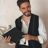 A man with a beard, wearing a white shirt and a Black Hand Embroidered Vest from AYA Sacred Wear, is sitting indoors and smiling while holding an open black book.