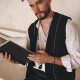 A man with a beard sits on a staircase, cradling a book. His black hand-embroidered vest from AYA Sacred Wear, crafted from organic cotton, complements his white shirt against the backdrop of a minimalist, light-toned wall.