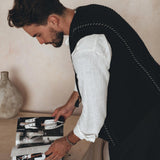 A person with short hair and a beard, wearing an AYA Sacred Wear Black Hand Embroidered Vest over a white shirt, is standing and flipping through a magazine. A beige vase sits on the table next to them against a neutral-toned wall.