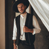A person with a beard stands indoors wearing a wide-brimmed hat, a white shirt, and the Black Hand Embroidered Vest for Men by AYA Sacred Wear. They hold a string of beads and stand next to a draped curtain.