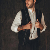 A bearded man dons a wide-brimmed hat, an AYA Sacred Wear Black Hand Embroidered Vest over a white shirt, and a long beaded necklace. He stands with a thoughtful expression against a dark, textured background, exuding an air of timeless elegance and natural style.