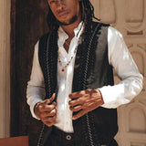 A person with long hair stands in front of a textured wall, wearing a white shirt and an AYA Sacred Wear Black Hand Embroidered Vest for Men that appears botanically dyed. The necklace adds flair to their relaxed expression as they adjust the handwoven vest. A wooden chair is partially visible beside them.