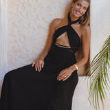 A woman wearing the Black Harmonia Dress by AYA Sacred Wear – an adjustable, gothic sexy dress also known as the Fairy of the River Dress – sits on a bench with a textured white wall behind her. She is smiling, has long hair, and is accessorized with large hoop earrings, a watch, and rings. A green plant is seen on the right side of the image.