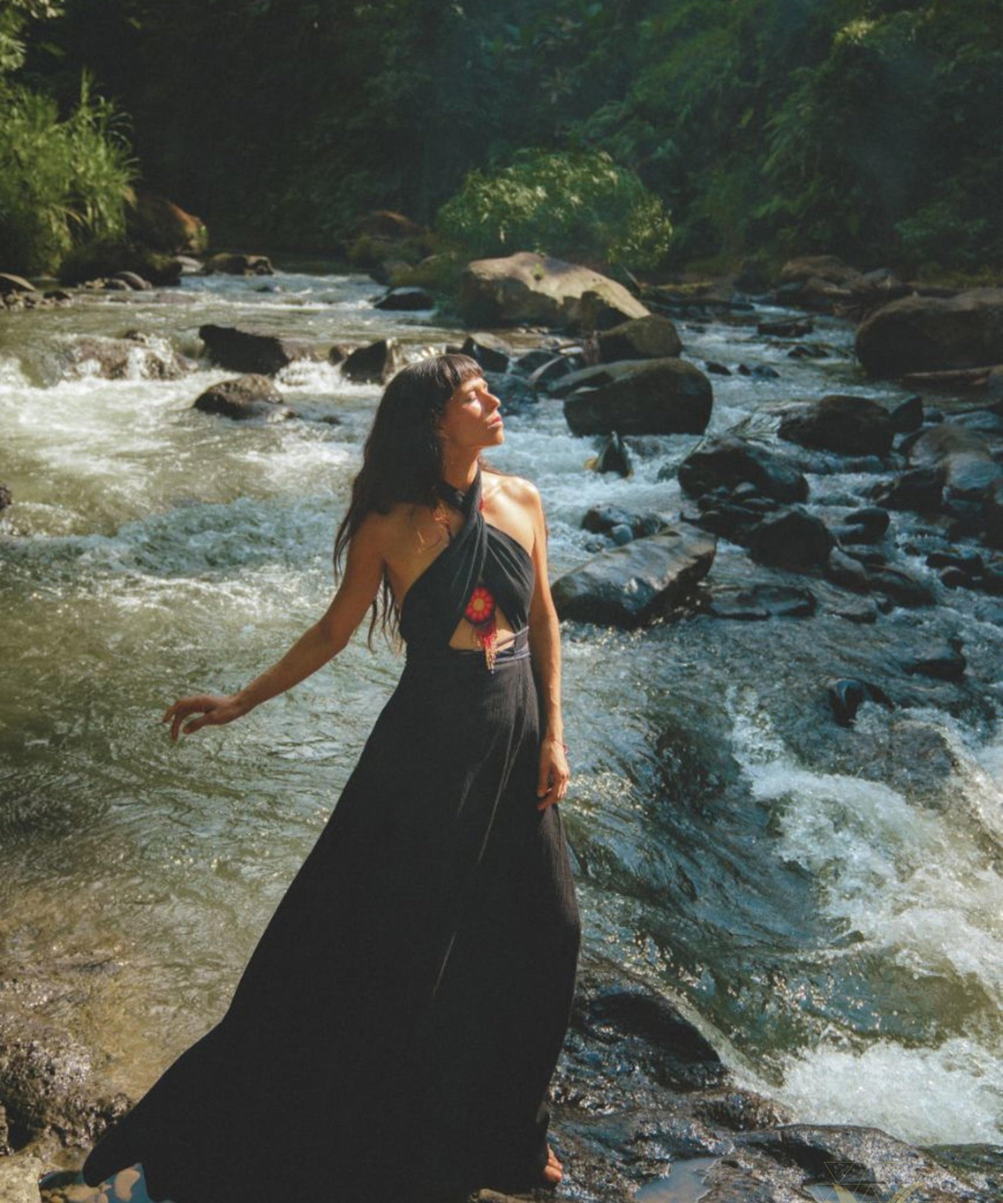 A woman stands on a rock in a flowing Black Harmonia Dress by the river. Sunlight filters through the trees, illuminating her face and the lush, green surroundings. The scenery, with its vibrant vegetation and flowing river, perfectly complements her dress—a blend of organic linen and cotton from AYA Sacred Wear's Gothic Sexy collection, also known as the Fairy of the River Dress.